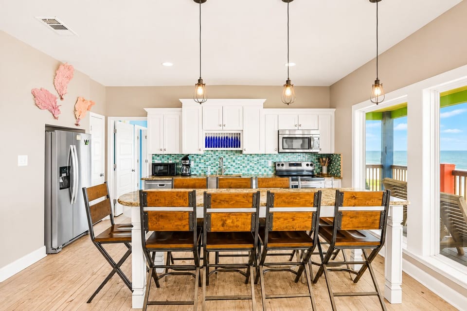Fully equipped coastal kitchen with granite breakfast bar, barstool seating, white cabinetry, stainless refrigerator and microwave, plus colorful tile backsplash.