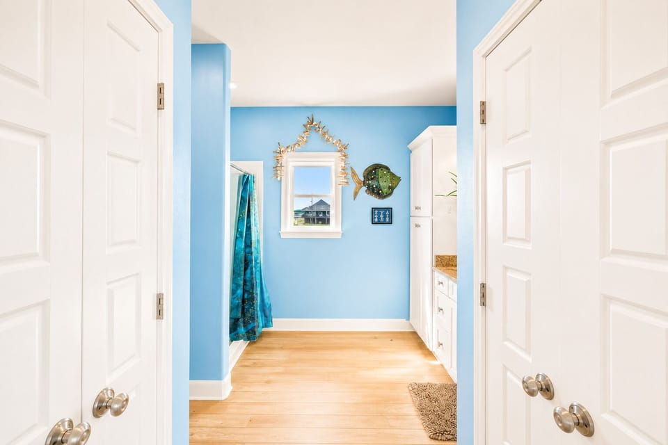Coastal hallway and vanity area with blue walls, wood-look flooring, decorative mirror and easy access between bedroom and bathroom spaces.
