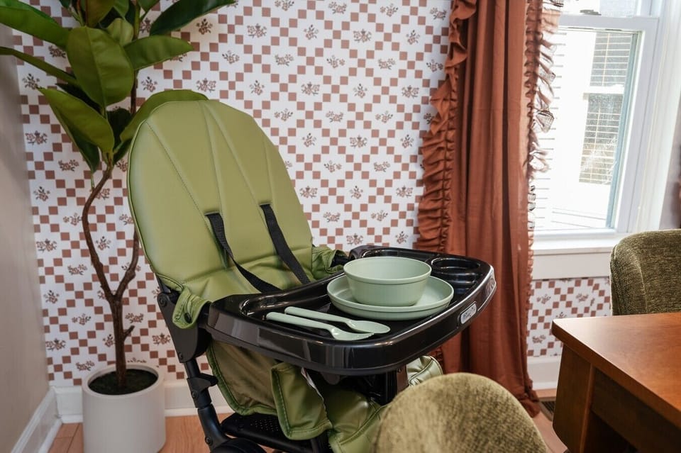 A charming feeding nook with a sage-green high chair and matching dishware, set against a playful vintage checkerboard wall. 🌿🥣