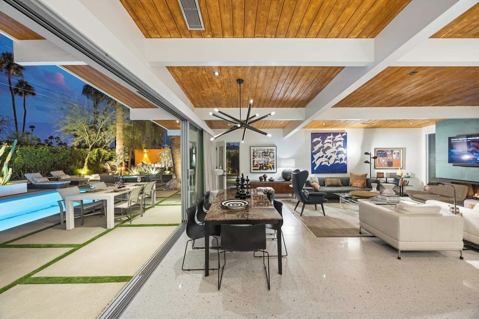 Open-air living room with vaulted wood ceilings and floor-to-ceiling glass walls dissolving into the illuminated pool beyond.