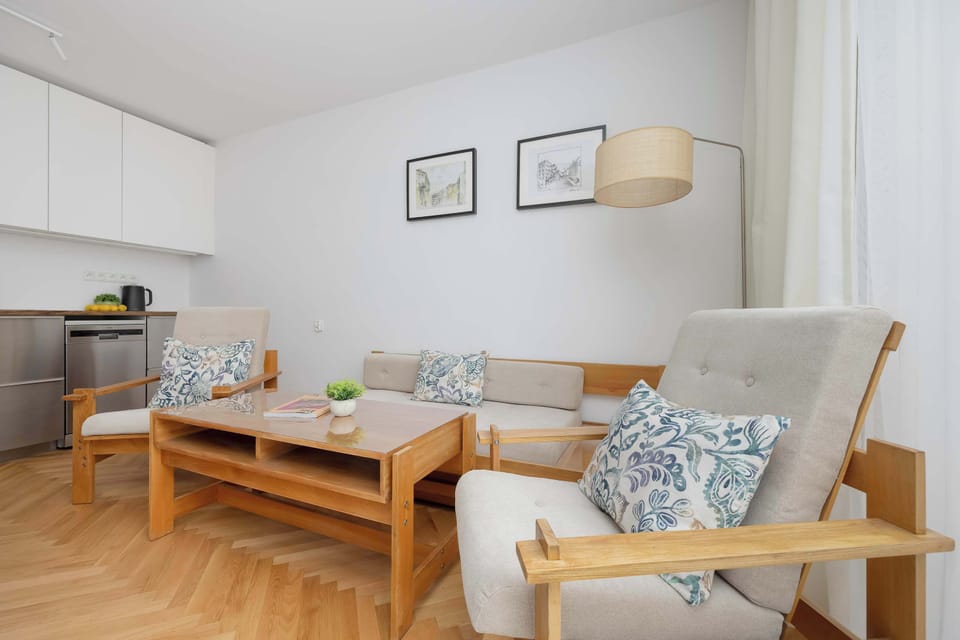 A cozy living room with a wooden coffee table, patterned armchair, and open kitchen in the background. Warm wooden flooring and soft textiles create a welcoming environment.