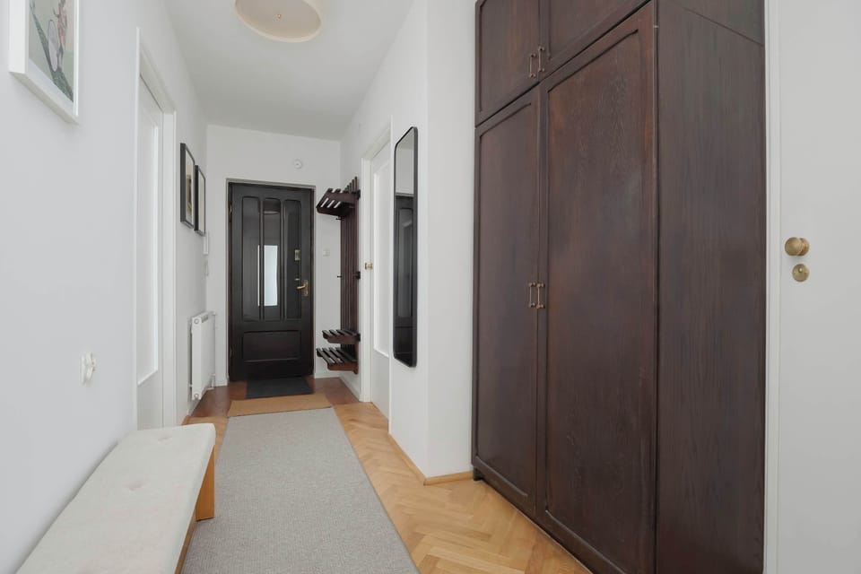 A long apartment hallway with wooden flooring, white walls, and dark wooden doors. The minimalist design creates a neat and organized entrance space.