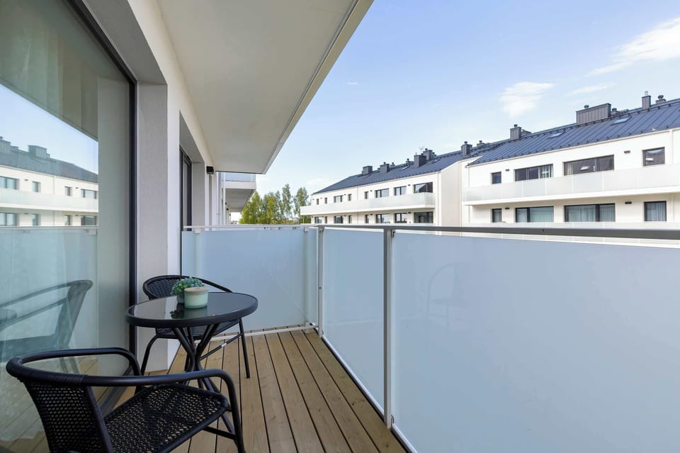 An outdoor balcony scene featuring a wooden deck floor and protective glass panels. Two black chairs and a small table offer a private spot for fresh air.