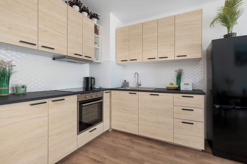 A modern kitchenette corner with light wood cabinetry and a contrasting black countertop. A sleek black refrigerator stands at the edge of the kitchen setup.
