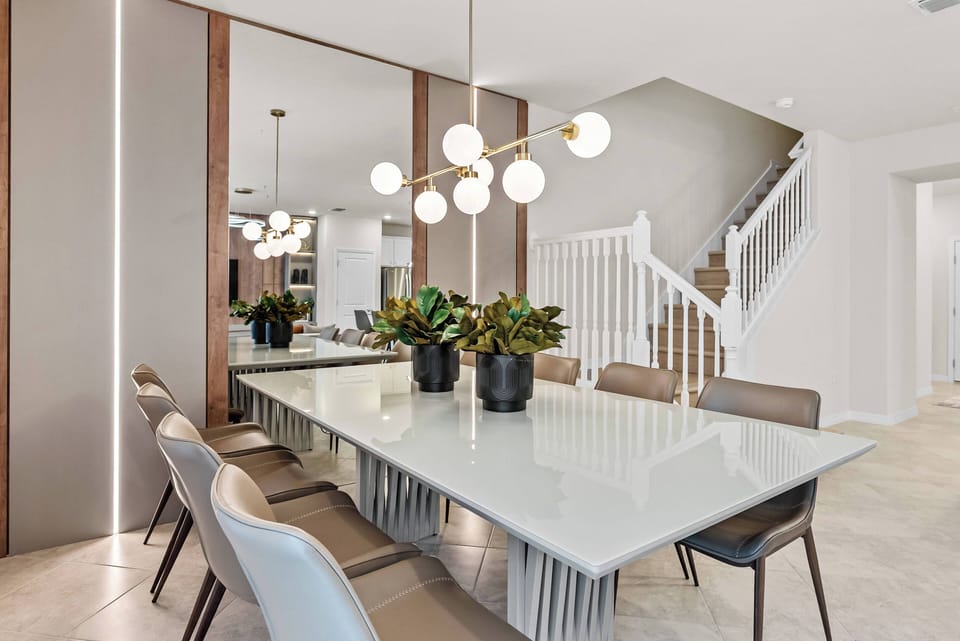 Savor your meal at the chic white dining table, surrounded by comfortable taupe chairs. The warm glow of the modern chandelier and refreshing greenery create an inviting atmosphere for a delightful dining experience.