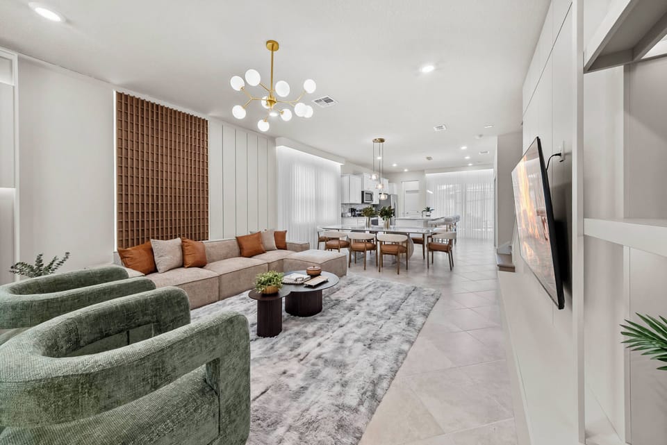 Experience a stylish living area with a plush green sofa and warm, earthy cushions. A modern chandelier softly illuminates the sleek dining table, creating an inviting space to gather and relax in comfort.