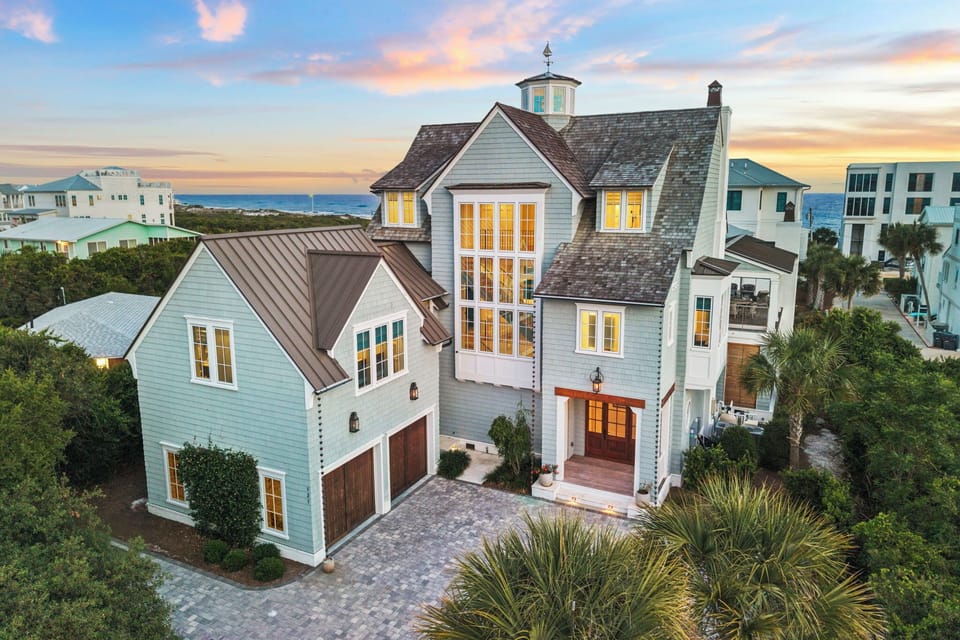 A charming coastal vacation home with distinctive architectural details sits in a peaceful beachside neighborhood, with ocean views and glowing windows welcoming guests as the day draws to a close.