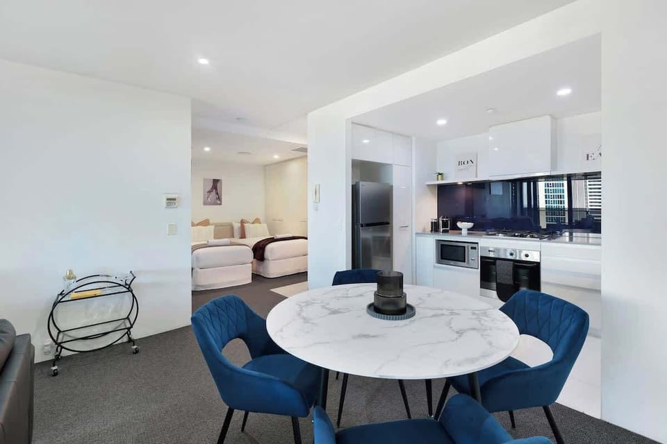 An elegant marble-finish dining setup transitions smoothly into a sleek, modern kitchen and a cozy bedroom alcove beyond. Perfectly maximizing space with premium finishes and a sophisticated palette.