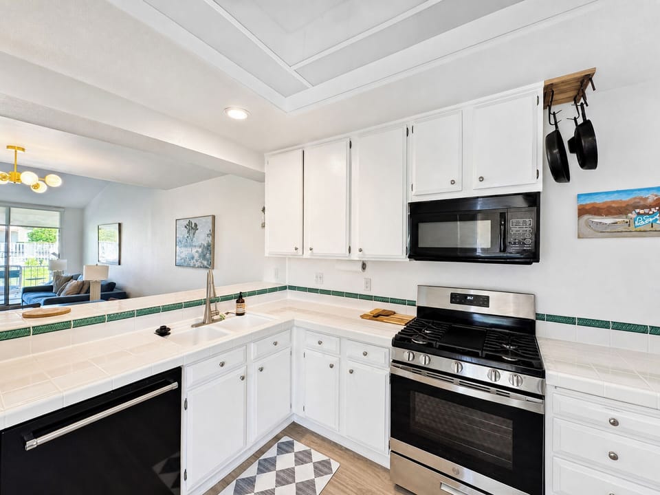 Your bright, fully equipped kitchen features crisp white cabinetry, cheerful tile accents, and modern appliances—ready for everything from morning coffee to home-cooked meals with ocean-view artwork overhead.