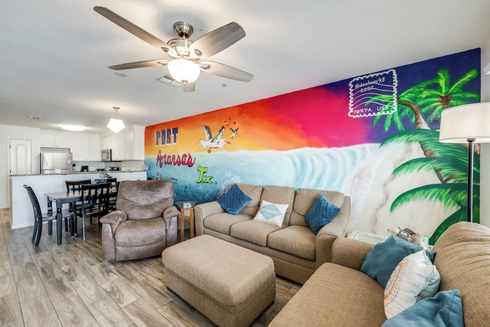 Unwind in your vibrant living space featuring a colorful Port Aransas beach mural, plush seating, and open-concept design connecting you to the fully equipped kitchen and dining area.