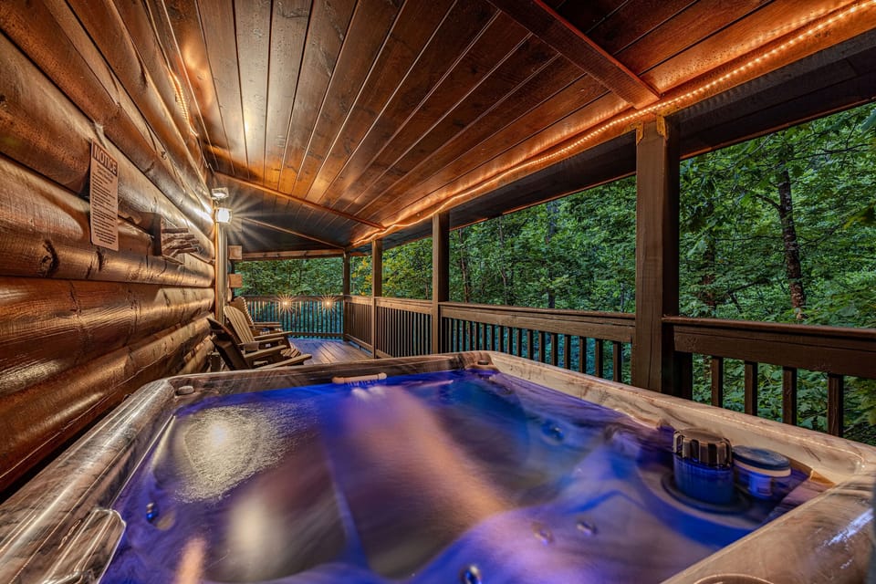 Unwind in the bubbling hot tub on the covered deck, where vibrant blue lighting creates a relaxing evening ambiance against the backdrop of the forest.