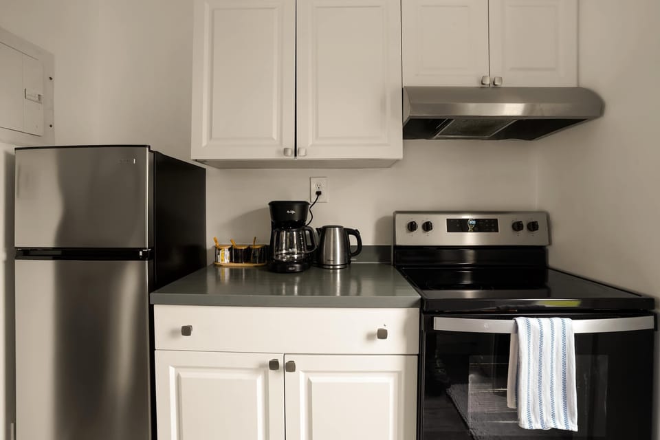 Full appliance lineup — stainless steel refrigerator, electric stove, coffee maker, and extractor hood. Fully stocked for cooking, baking, or simply making your morning coffee