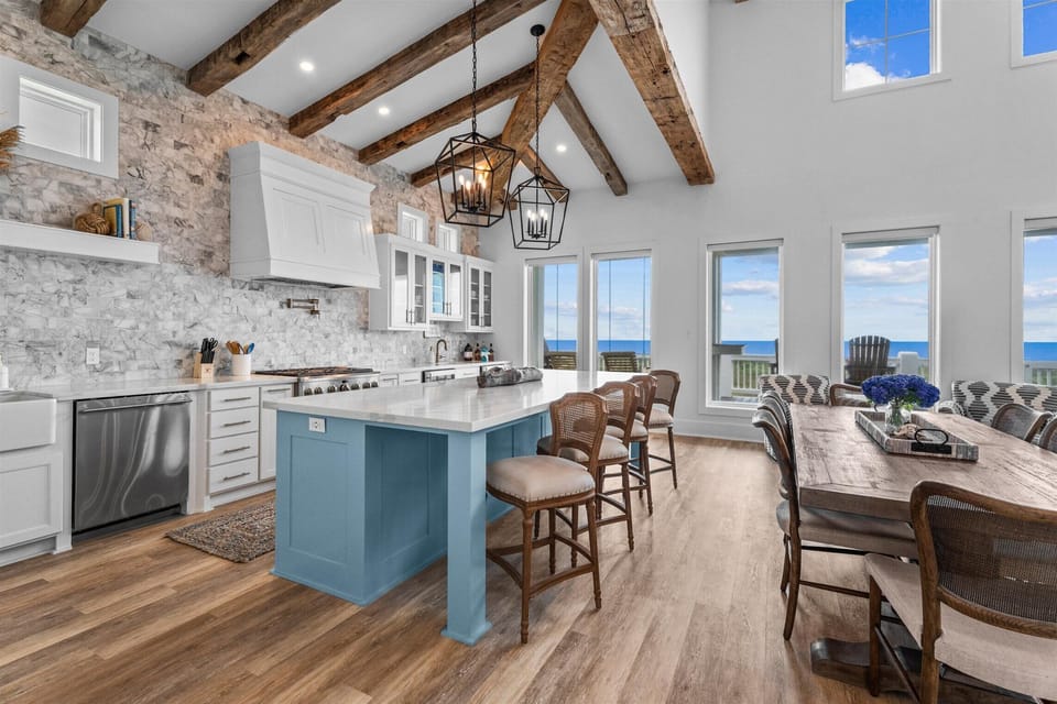 Luxury coastal kitchen with oversized island, stainless steel appliances, exposed wood beams, and open dining and living spaces.