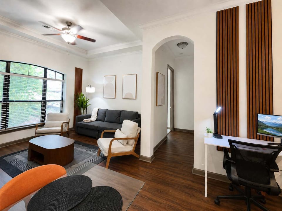 Bright, modern living room with high ceilings, wall-mounted TV, large arched window, dark sofa, accent chairs, and warm wood finishes.