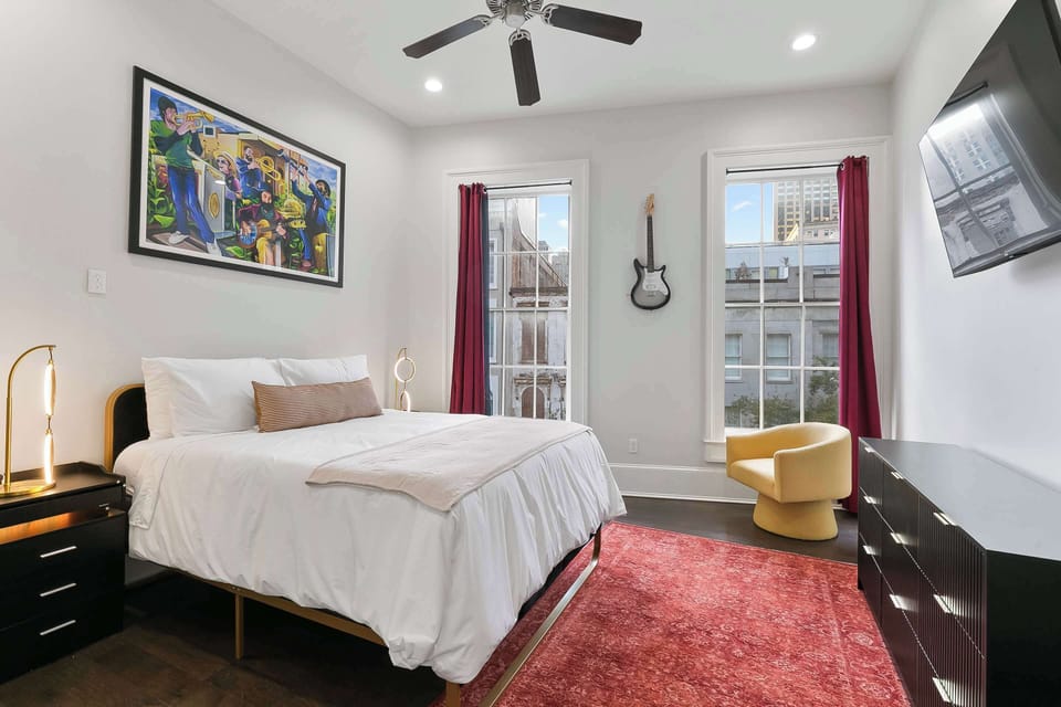 Bright art-filled suite showcasing a large Kalli Padgette painting and mounted guitar décor. Plush queen bed, smart TV, sunny windows, red rug, and modern comforts just minutes from Magazine Street and the French Quarter.