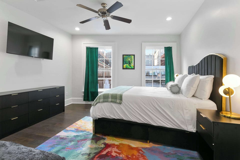 Charming bedroom showcasing wall art by Ayodele Owolabi. Queen bed with plush linens, smart TV, vibrant rug, large windows with city views, and generous closet space—perfect for art lovers near Magazine Street and the French Quarter.