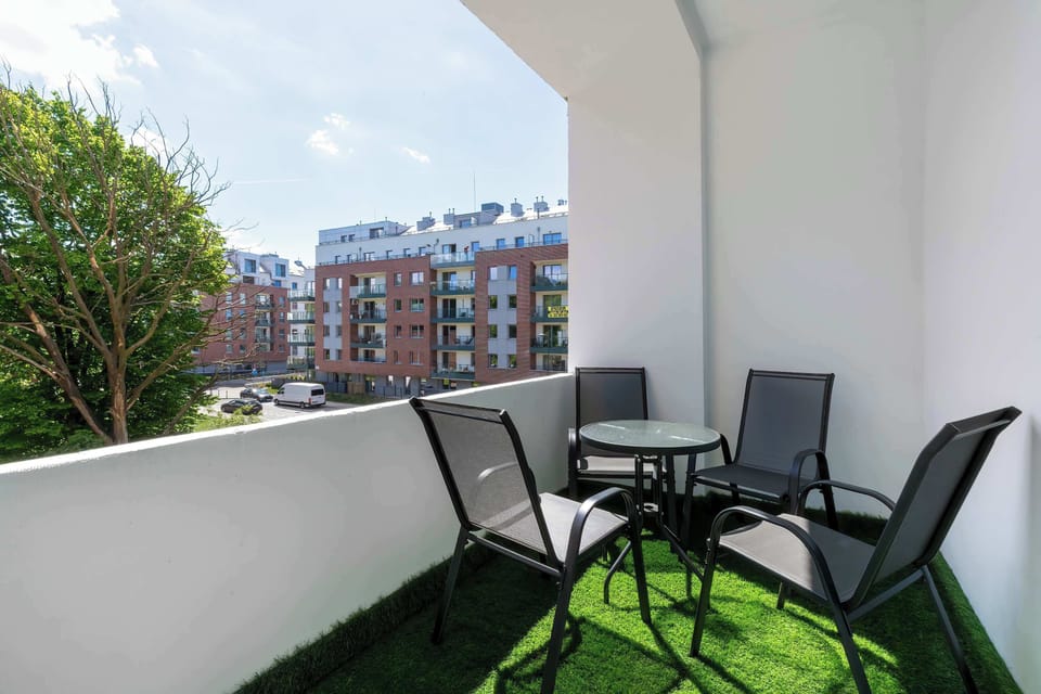 A balcony furnished with black outdoor chairs and a small table overlooking nearby residential buildings. Artificial grass flooring adds a cozy outdoor touch.