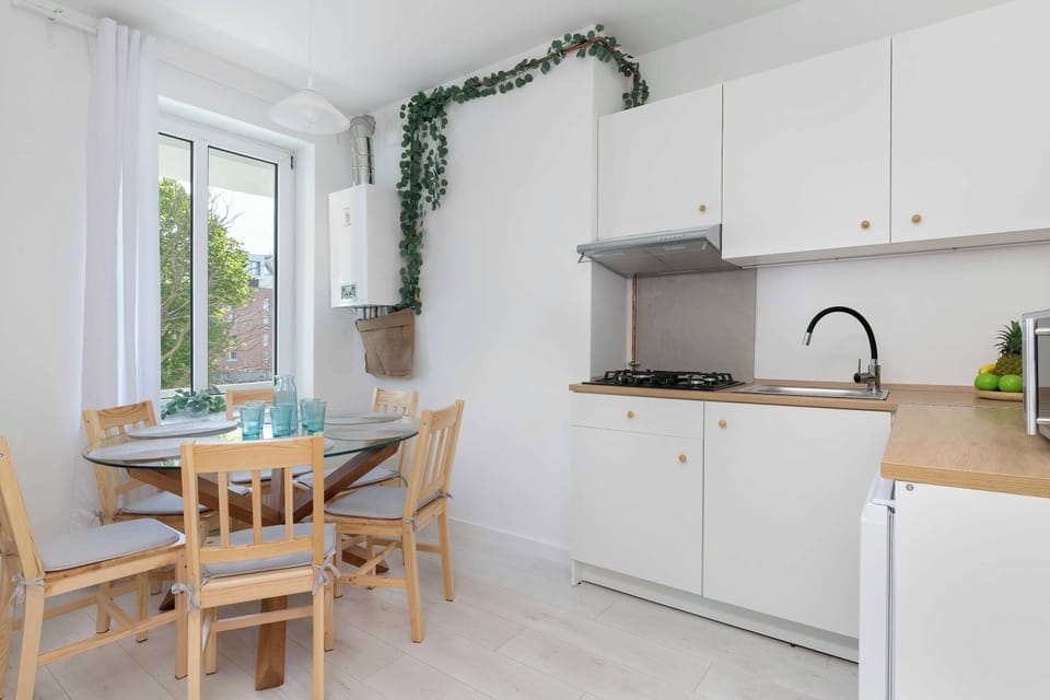 A small dining nook beside the kitchenette with wooden chairs and decorative greenery hanging on the wall. Light finishes and compact furnishings maximize the available space.