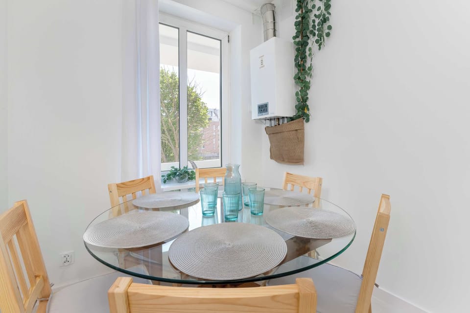 A charming dining table arranged beside large windows with simple tableware and decorative plants. The bright interior creates a pleasant spot for meals and coffee breaks.