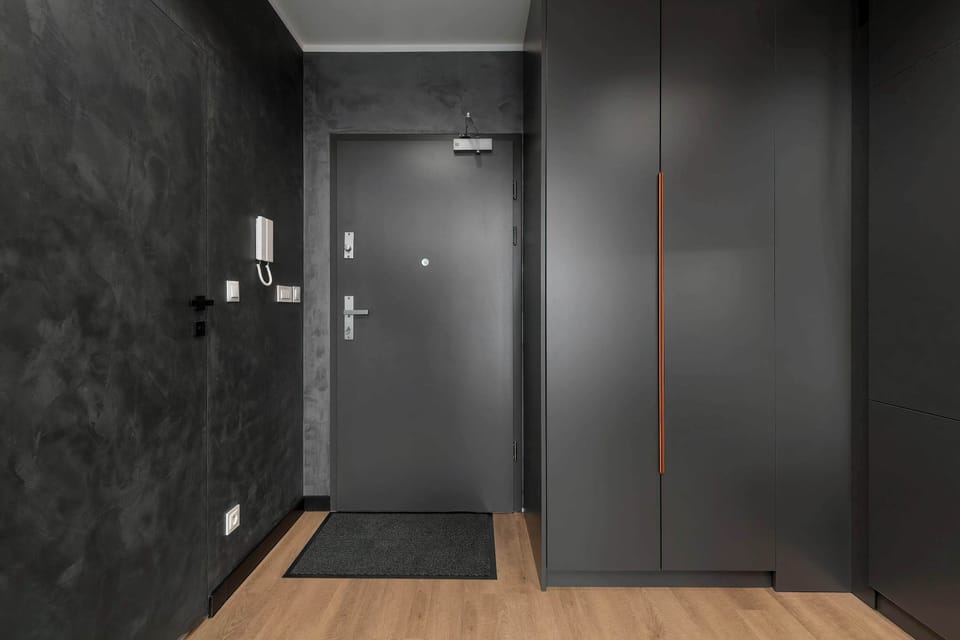 The apartment entrance is equipped with a dark grey front door and a spacious wardrobe featuring minimalist handles. A practical, dark doormat is laid on the floor right by the entrance.