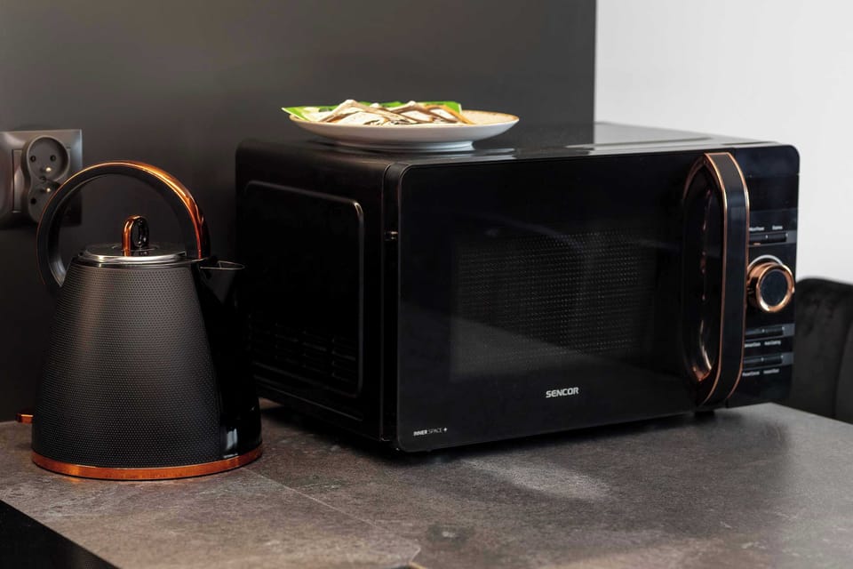 A black microwave oven with elegant gold accents and a matching electric kettle sit on the dark kitchen counter. A plate with a prepared coffee set is visible just above the appliances.