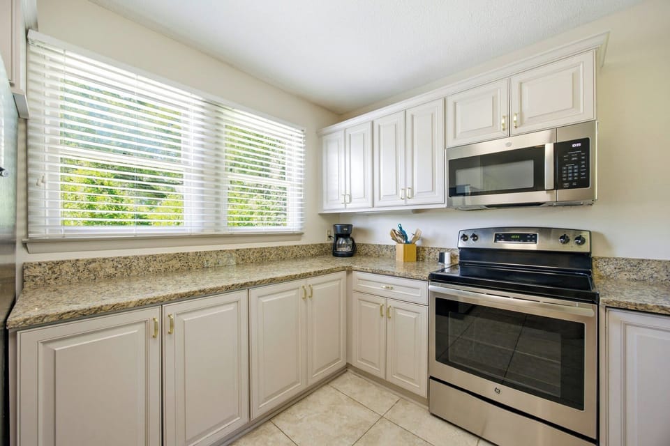 Speckled granite counters and sleek stainless appliances anchor this airy kitchen. Morning rays filter through the blinds, illuminating the clean white cabinetry and spacious prep areas for a cheerful start to your day.