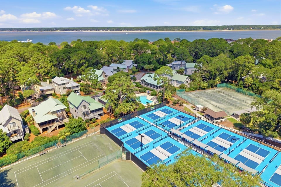 Vibrant blue pickleball courts and green tennis grounds sit amidst a canopy of pines, offering a playful escape just steps from the water. Afternoon sun illuminates this active community hub where sport meets the serenity of the shoreline.
