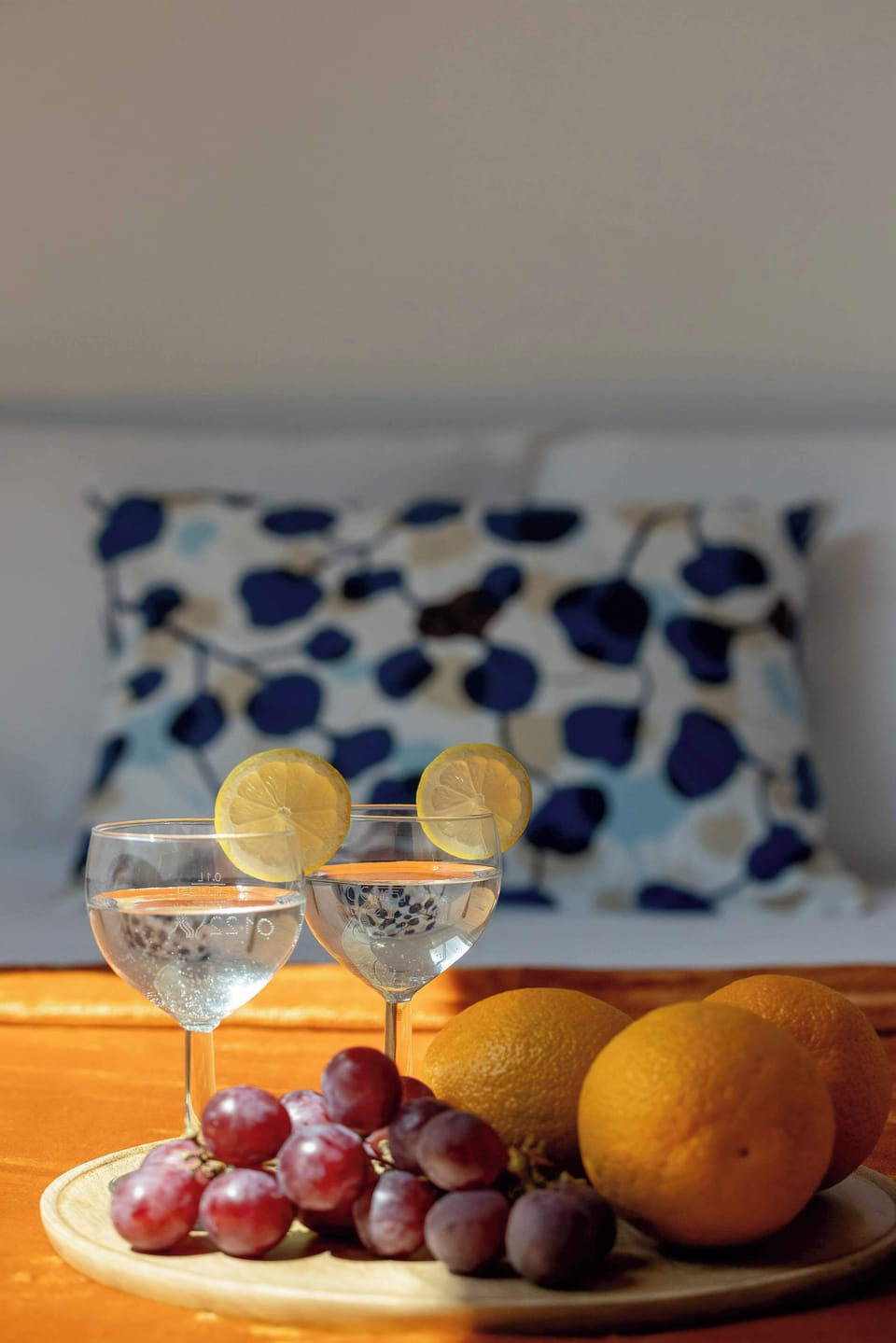 A detailed close-up shot features two wine glasses filled with a clear beverage and lemon slices, arranged next to a cluster of red grapes and oranges. The vibrant fruit arrangement sits on a wooden platter on top of the orange blanket.