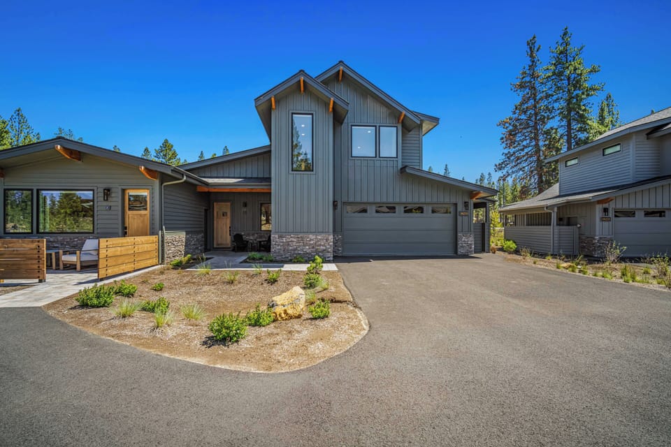 A striking modern mountain home welcomes you with a distinctive vaulted roofline, natural stone accents, and mature pines framing the entrance beneath a brilliant blue sky.