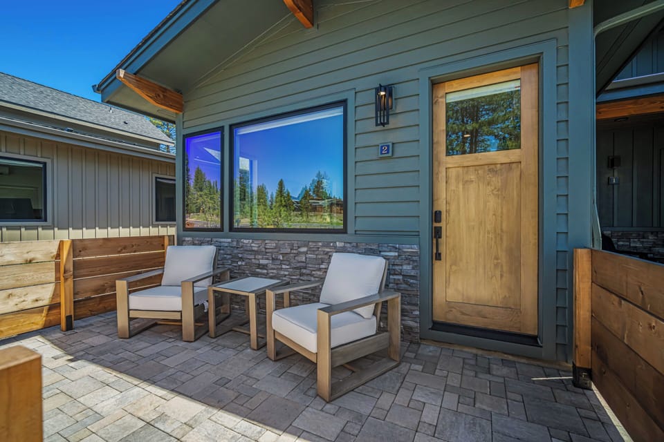 Modern vacation rental entrance with inviting patio seating under clear skies, surrounded by mountain scenery and evergreen trees. Comfortable outdoor area features contemporary furnishings and scenic views.