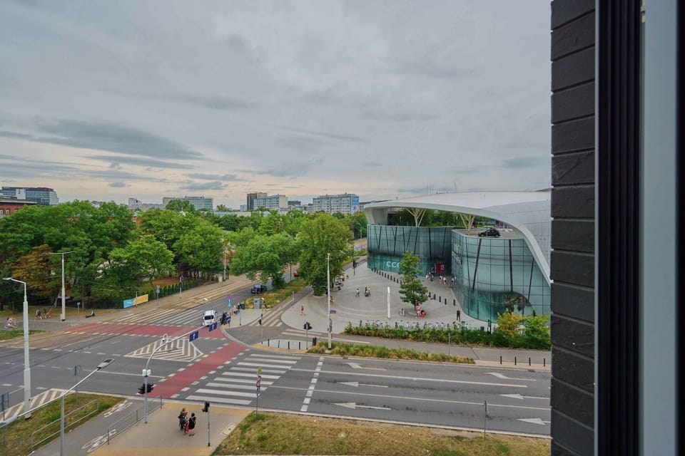 A city view with green trees, pedestrian crossings, and modern architecture in the background.

