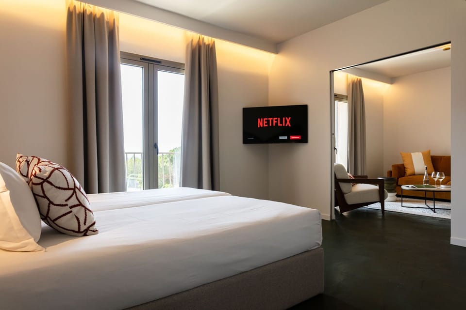 Two single beds facing a wall-mounted TV, with grey curtains framing the outdoor view