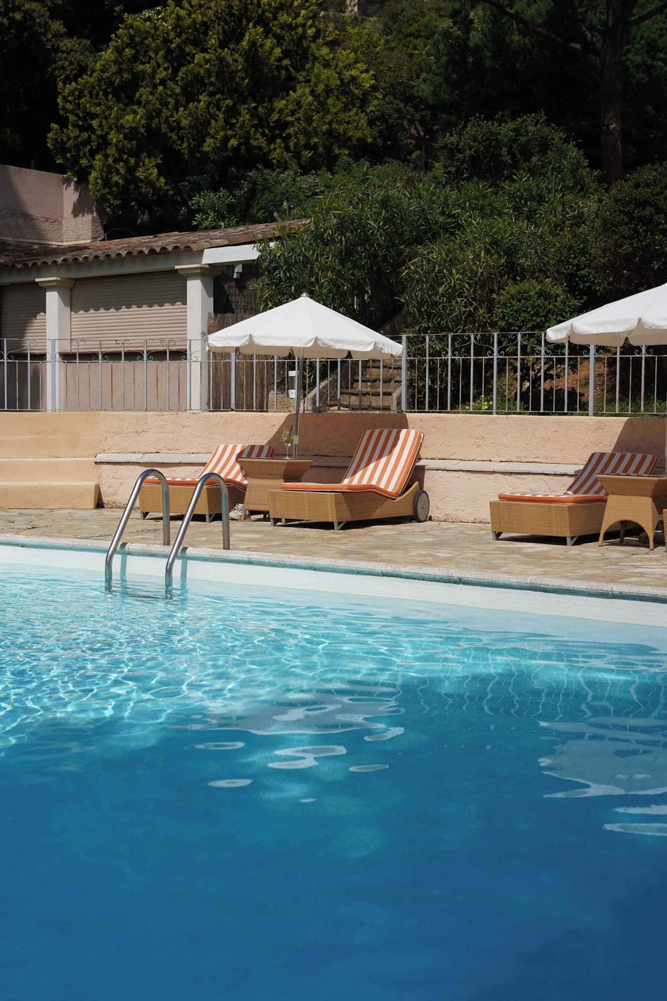 Blue pool water with striped lounge chairs and clipped hedges — afternoons here move slowly