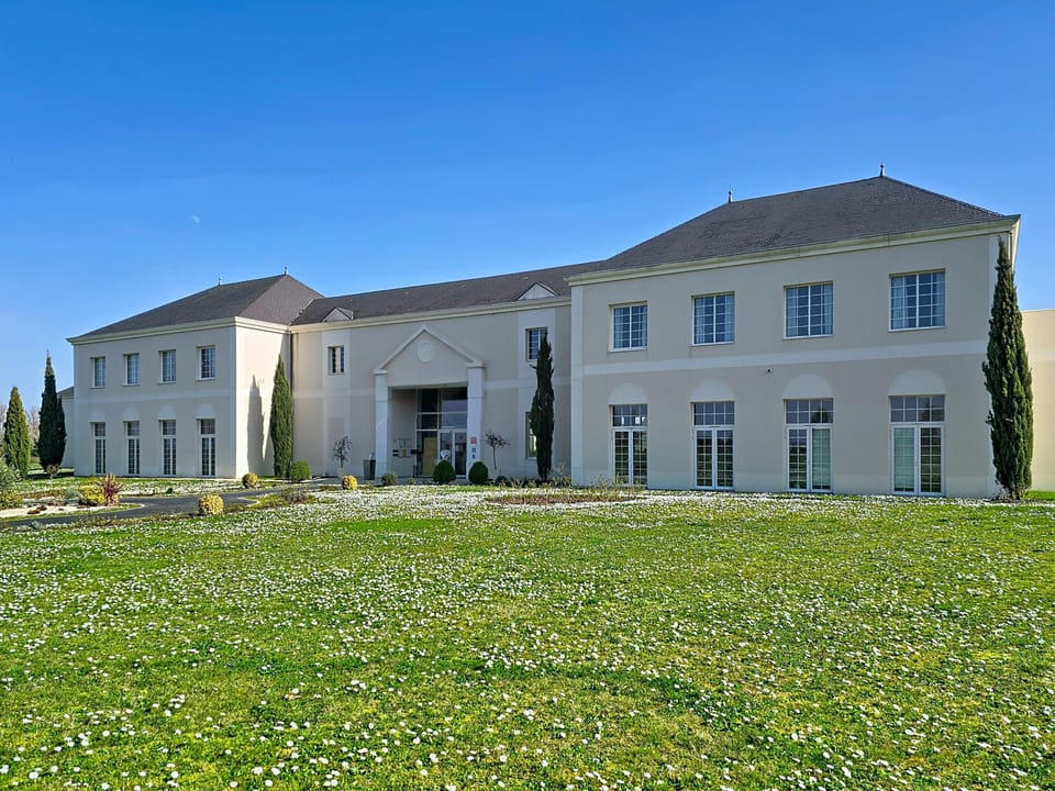 White-rendered two-storey building with tall windows and landscaped grounds planted with green shrubs