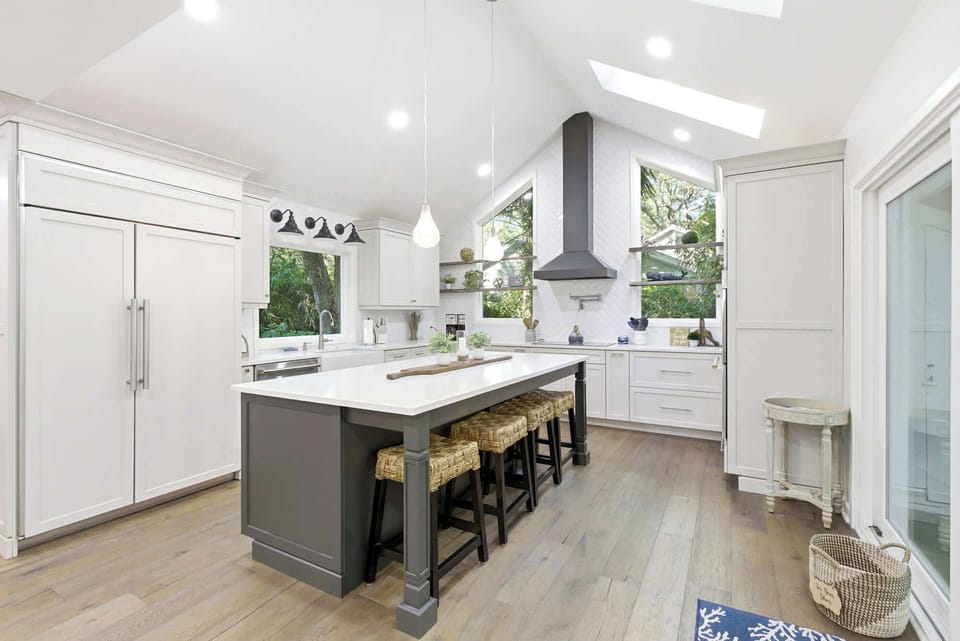 A chef's dream kitchen under vaulted skylights with white shaker cabinets, a gray island with wicker bar stools, pendant lights, open shelving with greenery, and a pool view through the slider.