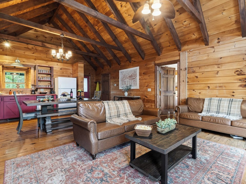 Cozy cabin living room with vaulted wood ceilings, a fireplace, and deck access surrounded by trees.