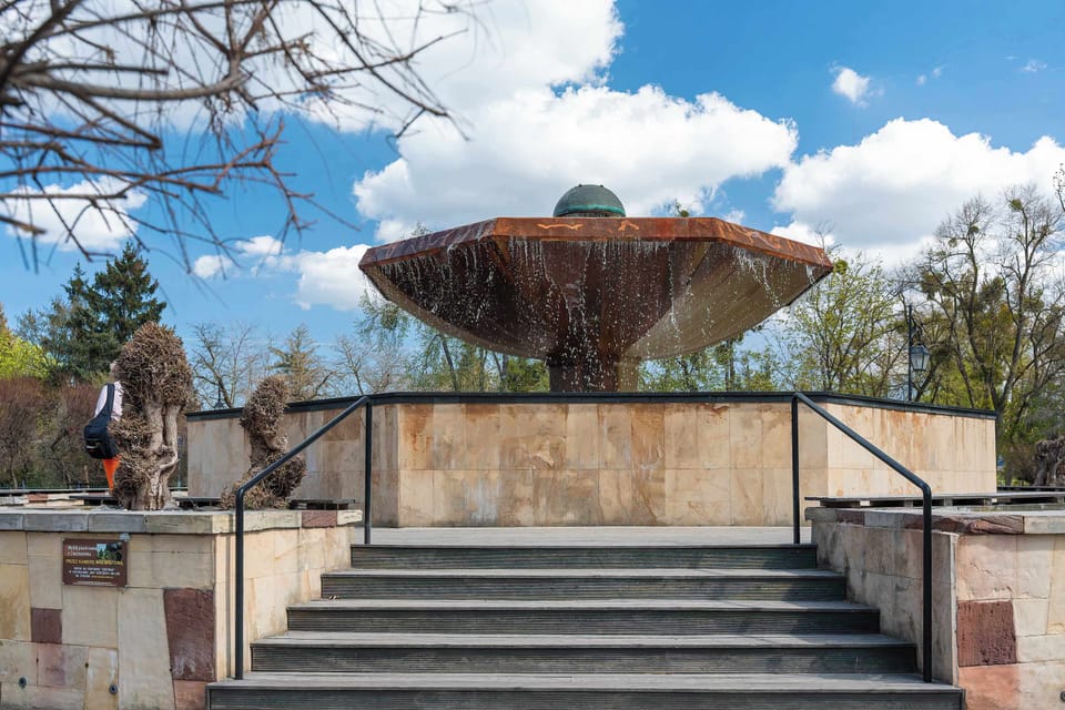 A multi-tiered stone fountain is the centerpiece of a public square, with water cascading over its large, flat basins. The structure is set against a bright sky with fluffy white clouds.