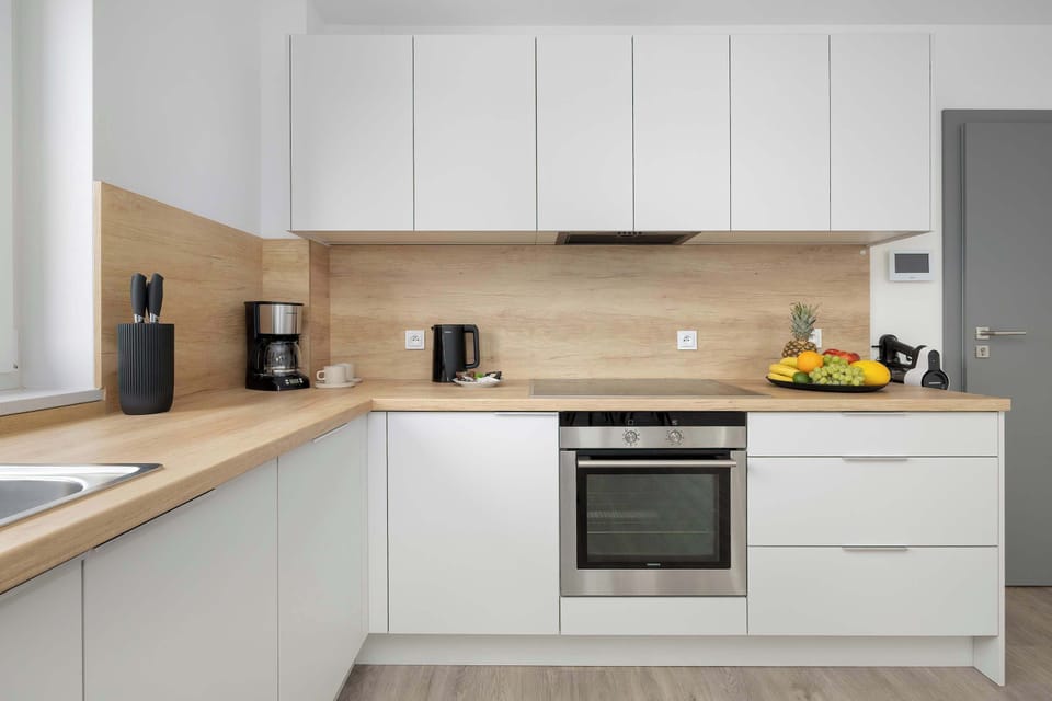 A modern L-shaped kitchen showcases crisp white cabinetry paired with a warm wooden countertop and matching backsplash. A built-in oven, stove, and integrated extractor hood make the space highly functional.