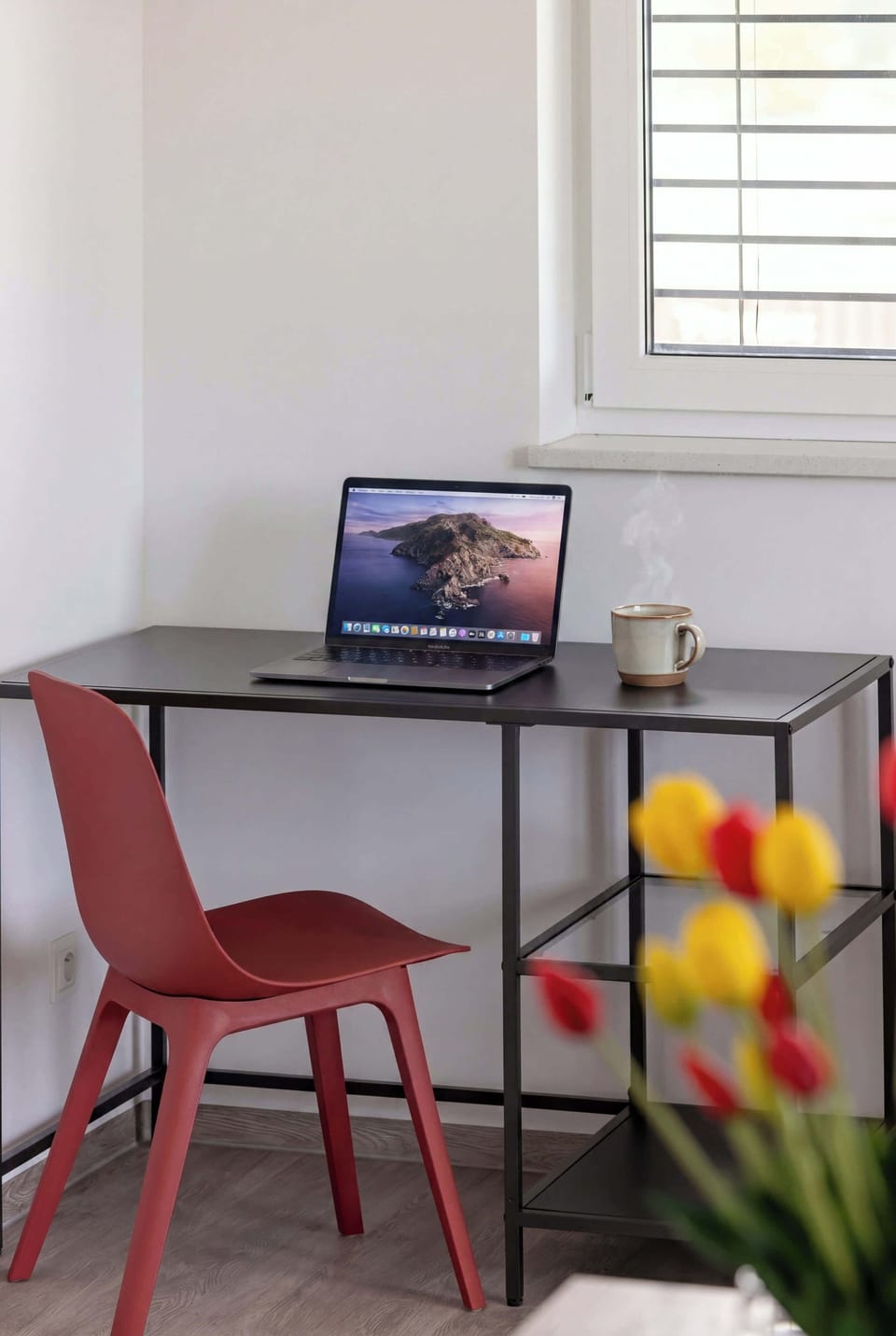 A vertical frame focuses on a minimalist home office corner featuring a slim black desk and a red chair. A laptop sits open on the desk next to a window that lets in soft natural light.