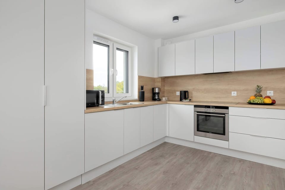 The minimalist white kitchen is captured from a side perspective, highlighting the clean alignment of the cabinetry. A large window above the countertop area ensures plenty of daylight during meal preparation.