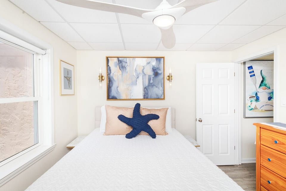 Unwind in your coastal-inspired bedroom with crisp white linens, calming ocean artwork, and a playful starfish accent pillow welcoming your beach getaway dreams.