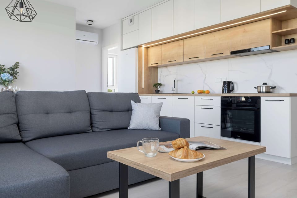 A contemporary living room connected to a kitchenette, furnished with a gray sectional couch and wooden coffee table. Minimalist décor and soft lighting create a welcoming ambiance.