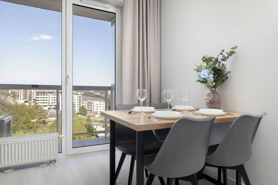 A dining corner beside floor-to-ceiling windows featuring a dark dining table and upholstered chairs. Panoramic urban views add elegance to the modern interior.