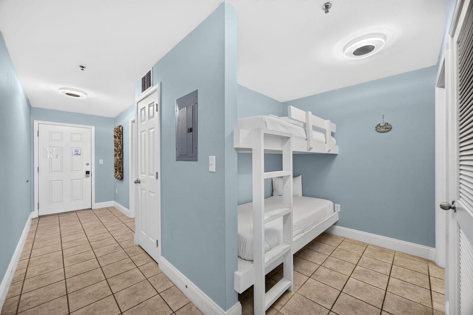 Your beach getaway features coastal-inspired spaces like this cheerful room with a white bunk bed, perfect for kids to share adventures and drift off after a sun-soaked day.