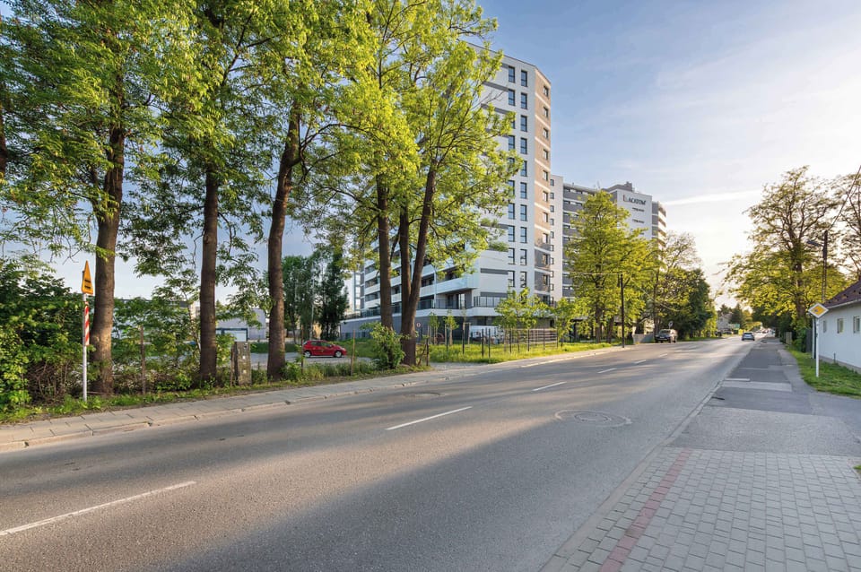 A street view lined with trees and modern apartment buildings. The environment feels calm, residential, and well planned.