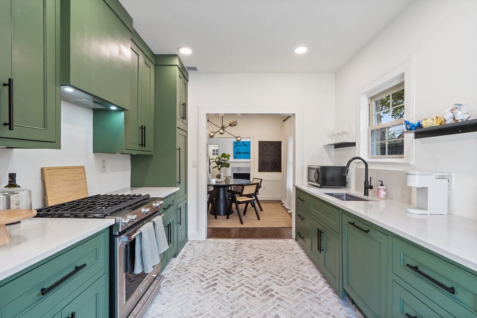 Kitchen with sage green cabinetry, gas range, and open view to dining area. Fully stocked with cookware, dishes, silverware, cooking basics, wine glasses, and Keurig coffee maker. Dishwasher included.