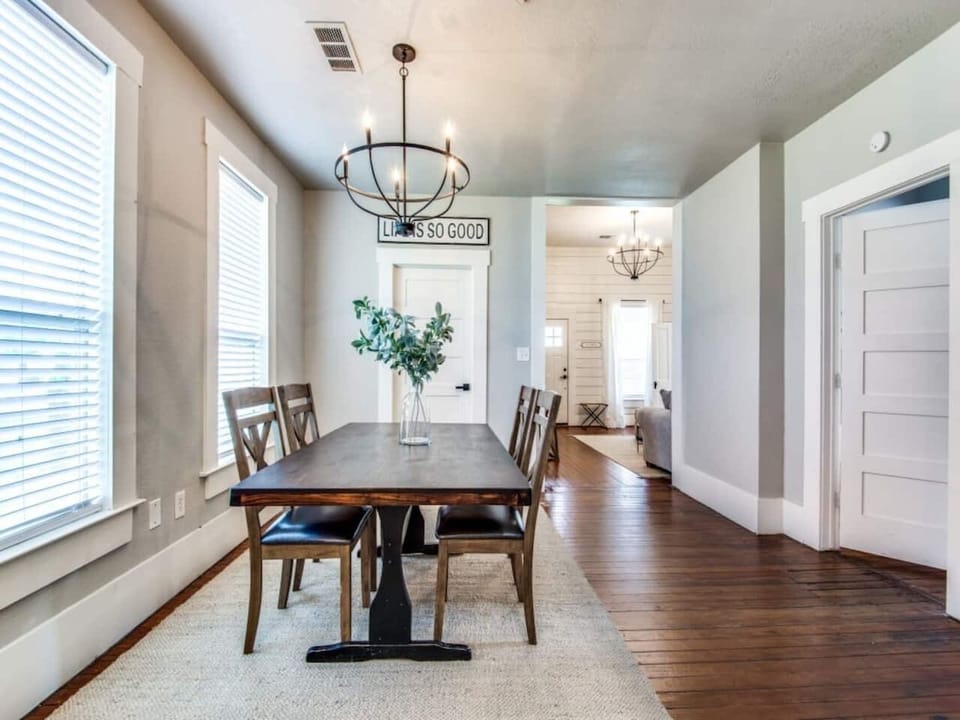 Farmhouse dining table with upholstered chairs, statement chandelier, shiplap accent wall, and hardwood floors. Bright natural light from large windows. Modern farmhouse Airbnb in downtown Aubrey TX steps from S Main Street dining.