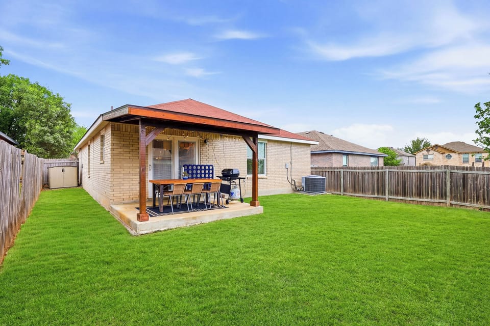 Spacious covered patio and open green space with string lights, outdoor dining set, and propane BBQ grill — all set against a fully fenced, private backyard with lush green lawn. Perfect for cookouts and evenings under the Texas sky.