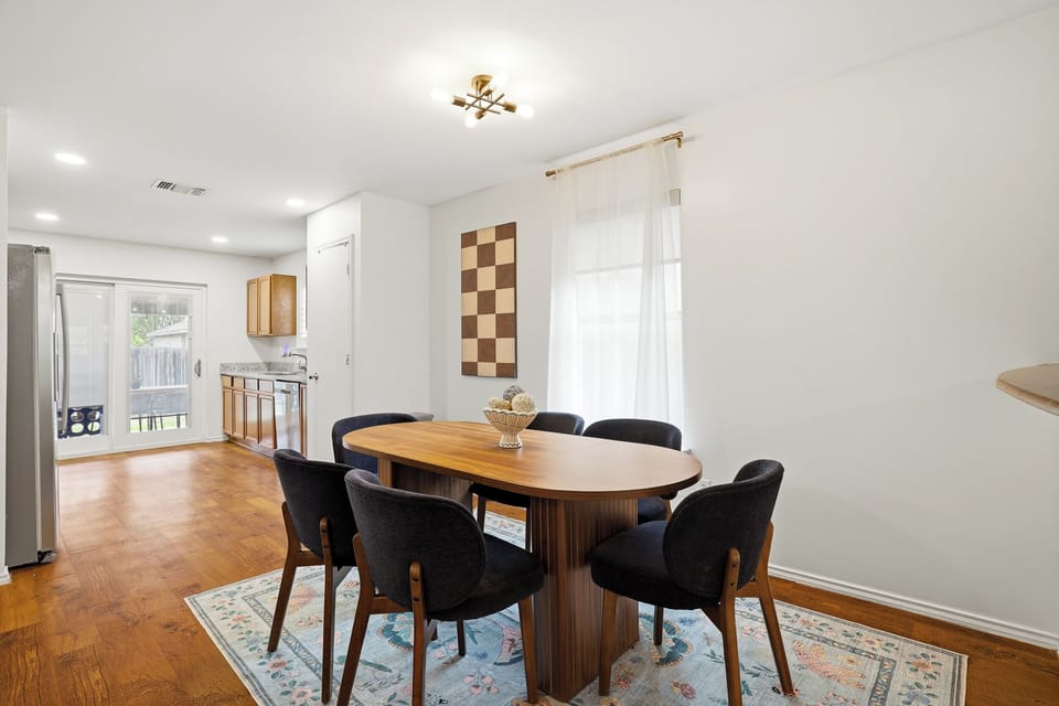 Bright dining room with seating for 6, gold pendant lighting, hardwood floors, and open sightlines through to the living room and front entry. Airy and well-lit for morning coffee or evening dinners.