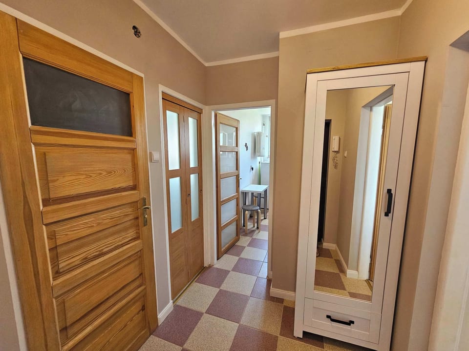 A welcoming entrance hallway showcases a traditional wooden front door and a tall white wardrobe with a built-in mirror. Checkered floor tiles lead further into the brightly lit rooms of the apartment.